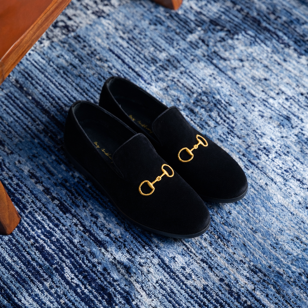 Black velvet loafers with gold horsebit detail on a blue textured rug